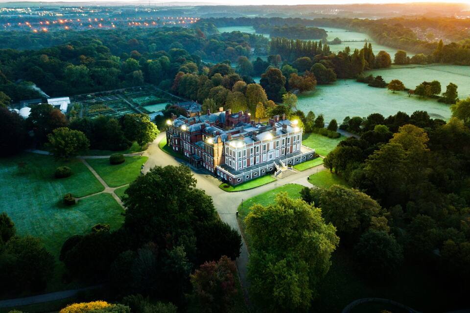 A large, historic building with multiple stories, surrounded by lush greenery and a river in the background.