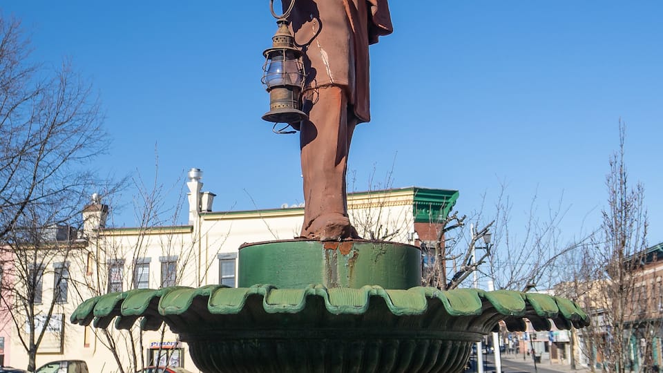 A statue of a man in a hat holding a lantern, standing on a green pedestal.