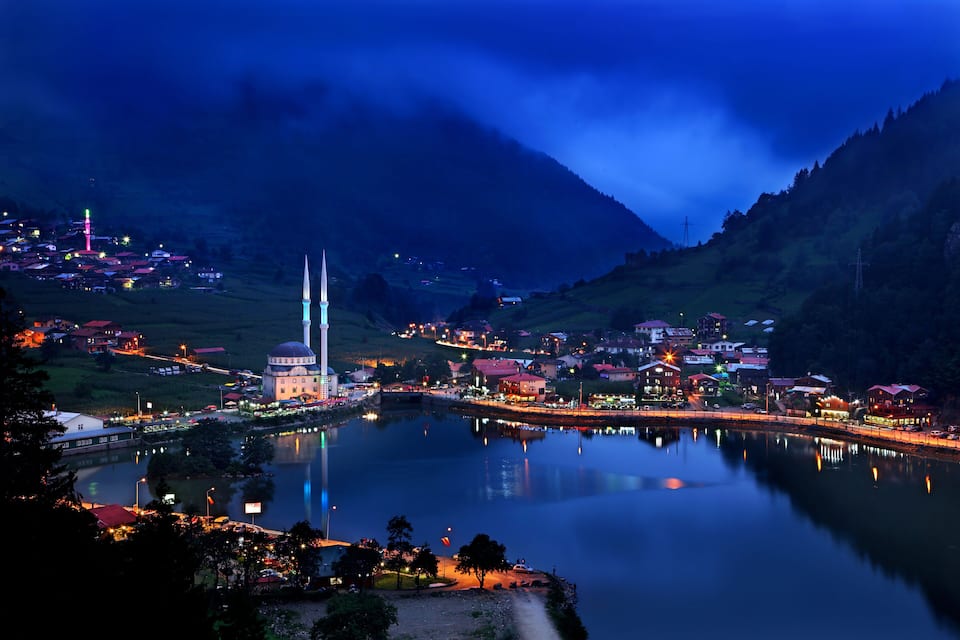 A town by a lake at dusk with illuminated buildings and a prominent tower.