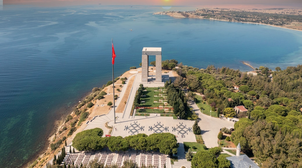 An aerial view of a coastal memorial with a flagpole, a monument, and a landscaped area with trees and pathways.