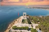 An aerial view of a coastal memorial with a flagpole, a monument, and a landscaped area with trees and pathways.
