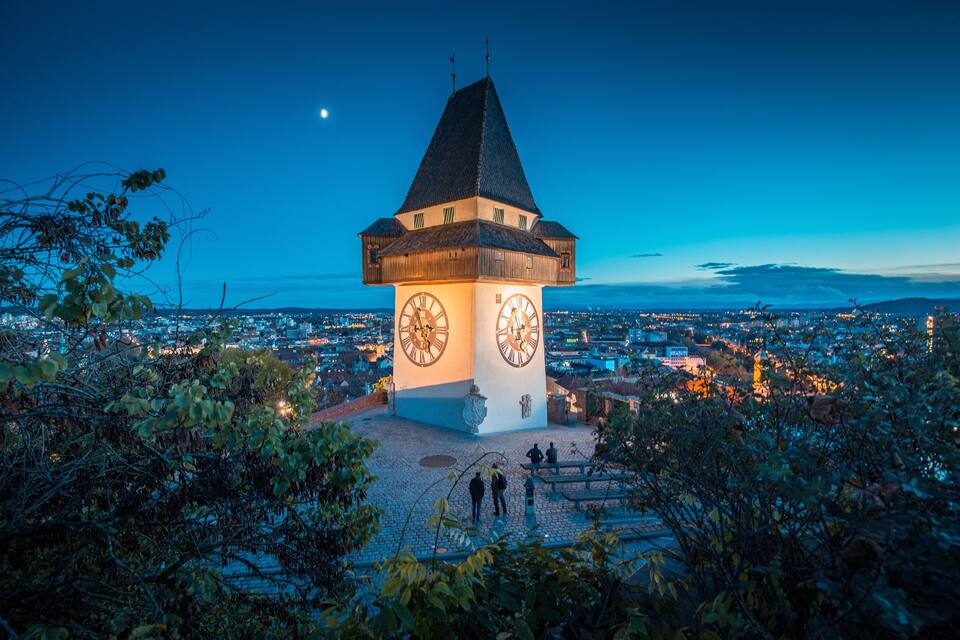A clock tower with two large clocks, surrounded by trees and overlooking a cityscape at dusk.