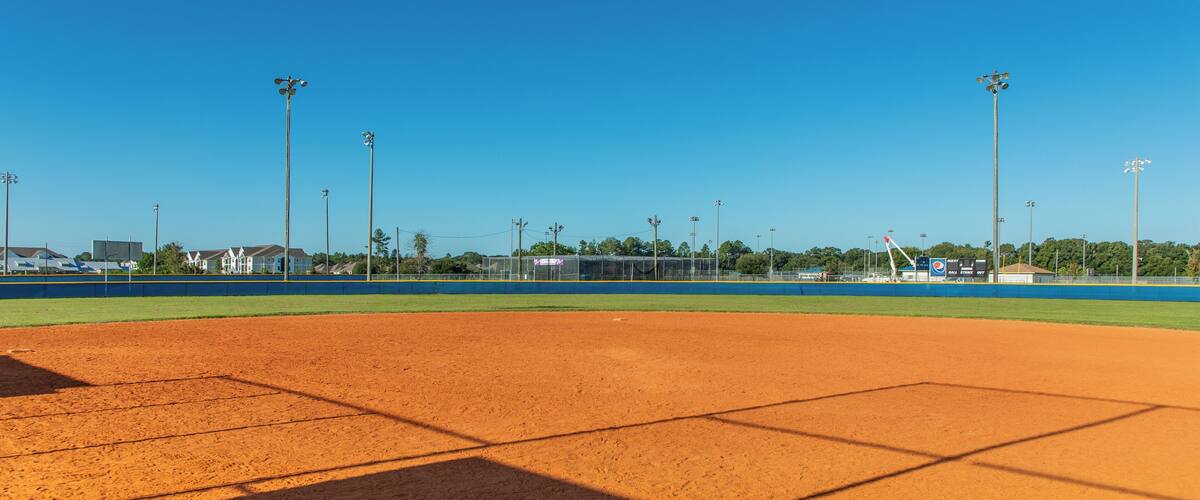 A baseball field with a clear blue sky, floodlights, and a scoreboard in the background.