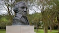 A bronze bust of a historical figure on a pedestal labeled "WASHINGTON" in a park with trees and grass.