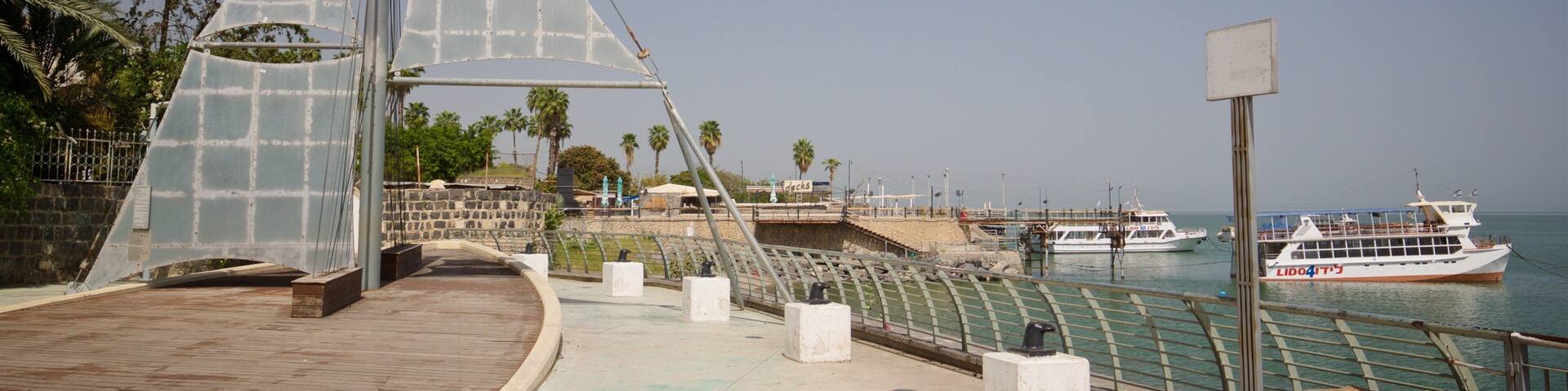 A coastal promenade with a modern glass bridge structure, a stone wall, and a view of a boat named LIDOTI.