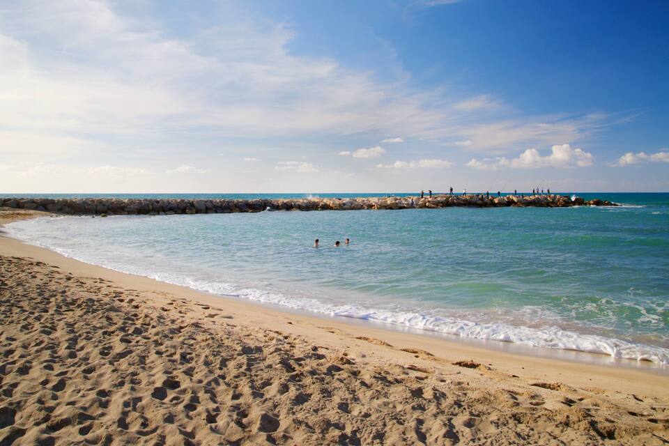 Una platja sorrenca amb aigües tranquil·les, un moll de pedra i un cel serè.