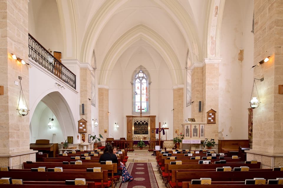 Interior de iglesia con altos techos abovedados, bancos de madera, un pasillo central y una vidriera.
