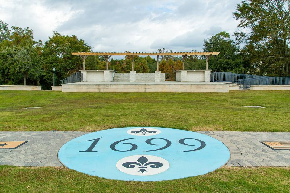 Un parque con un pabellón, un banco de piedra y un pavimento circular con los símbolos '1699' y fleur-de-lis.