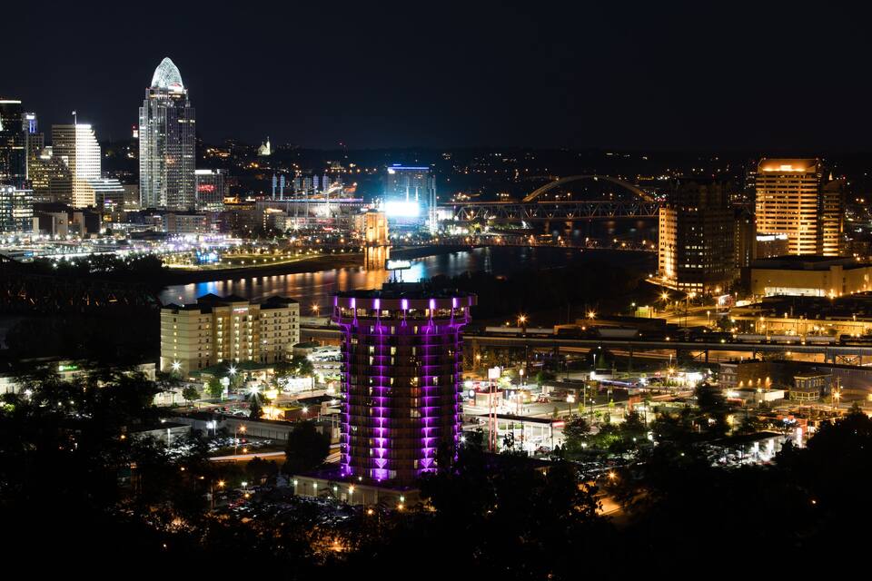 A cityscape at night with illuminated buildings, a prominent hotel with purple lighting, and a bridge in the background.