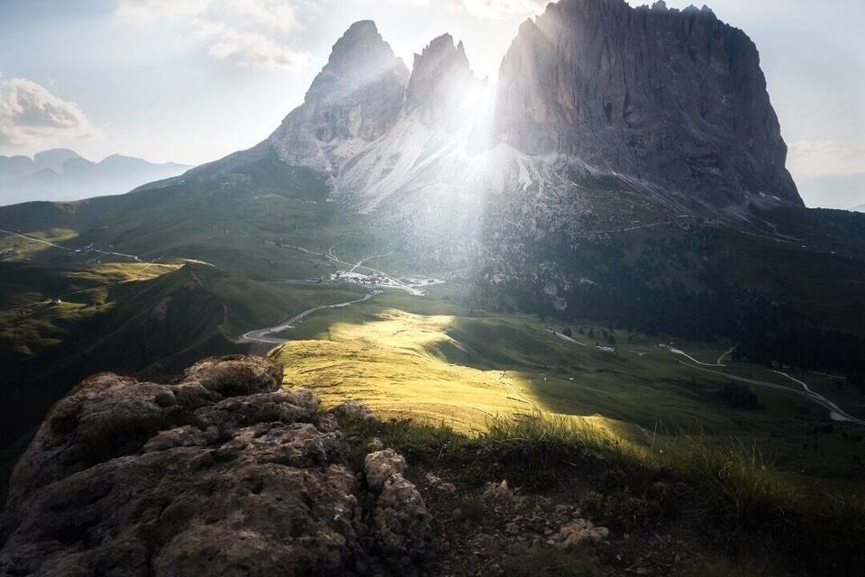 Un paisaje montañoso con un pico destacado, un valle con una carretera serpenteante y un primer plano rocoso.