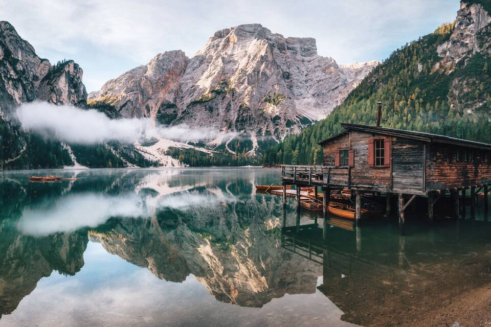 A wooden cabin on stilts by a calm lake with mountains in the background.