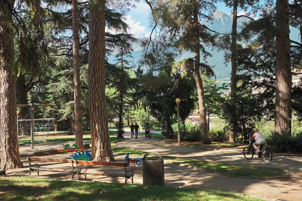 Um parque com árvores altas, um parquinho e um ciclista.