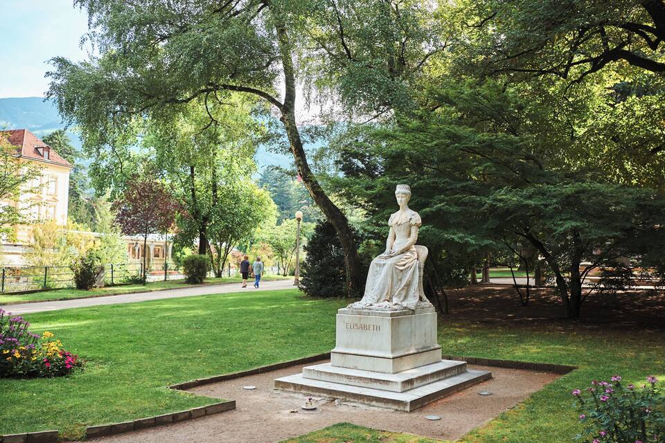 Una estatua de una mujer llamada Elisabeth en un parque con árboles y un edificio al fondo.