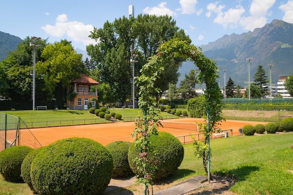 Un court de tennis entouré de pelouses bien entretenues, d’un petit bâtiment et, au loin, de montagnes.