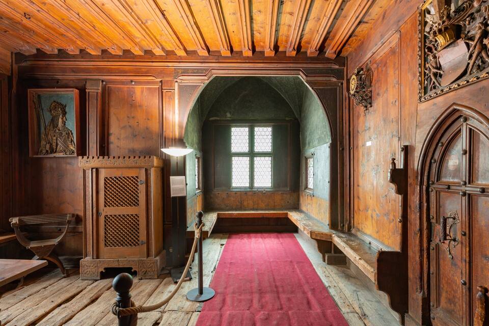 A wooden interior with a red carpet, wooden benches, and a large arched window.
