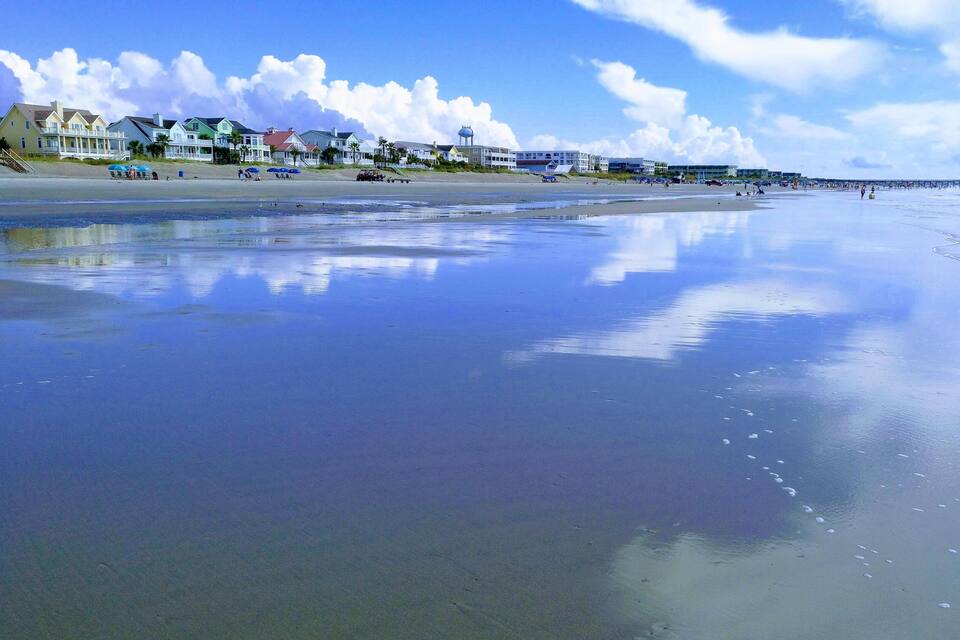 En strand med en spejlblank vandoverflade, huse og en mole i baggrunden.