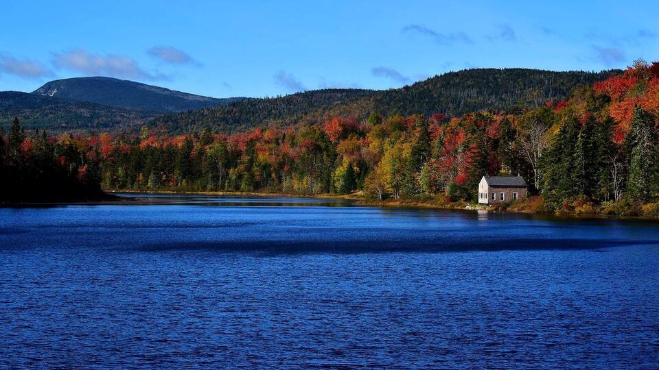 A lake with a house on the shore, surrounded by autumn-colored trees and a mountain in the background.