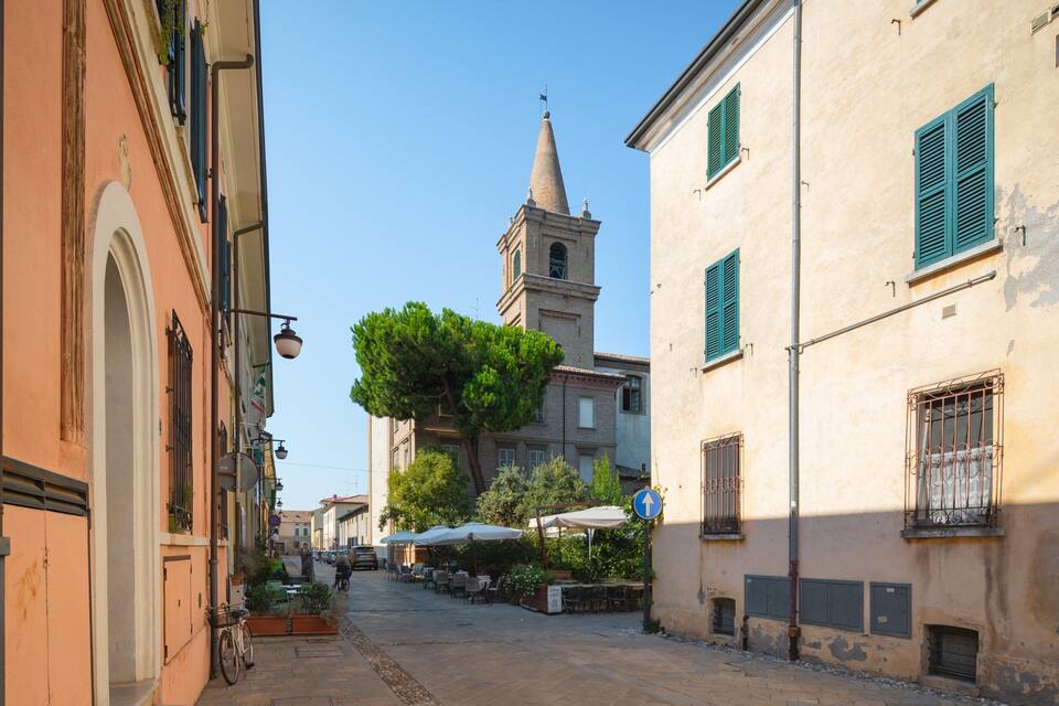 Una stretta via fiancheggiata da edifici, con una torre campanaria e tavoli all'aperto.