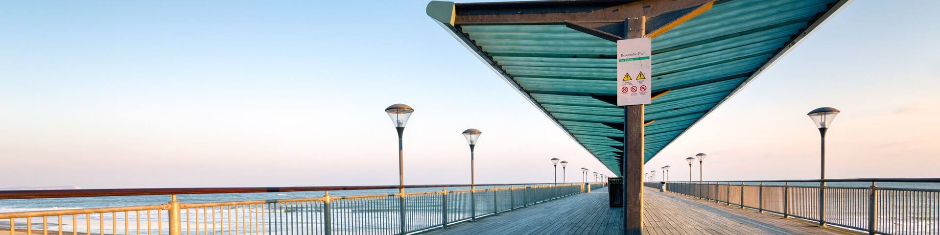 A wooden boardwalk with a metal railing, street lamps, and a sign indicating a park entrance.