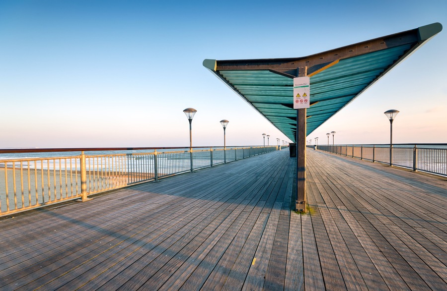 A wooden boardwalk with a metal railing, street lamps, and a sign indicating a park entrance.