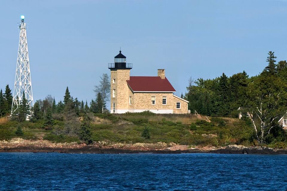 Un faro en una isla rocosa rodeada de un bosque denso y un cielo azul despejado.