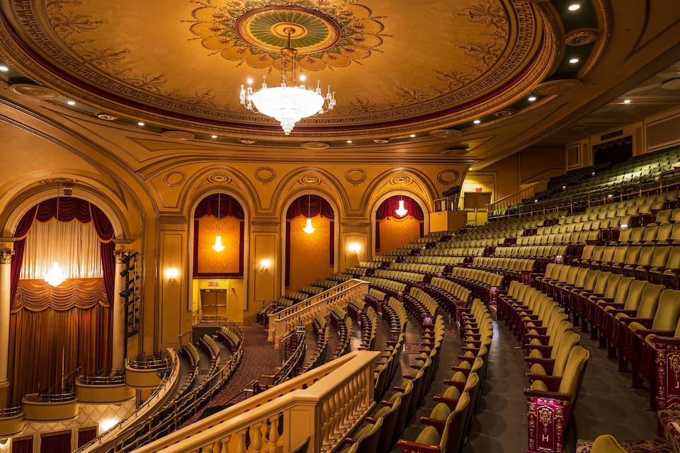 A grand theater with ornate ceiling, rows of plush seats, and a chandelier.