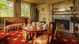 A room with a floral sofa, a round wooden table with tea sets, a fireplace, and a large window with greenery outside.