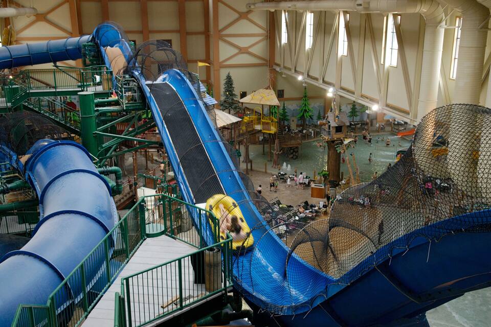 Parque acuático interior con un tobogán de agua azul grande, una zona de juegos con redes y una terraza de madera con gente.