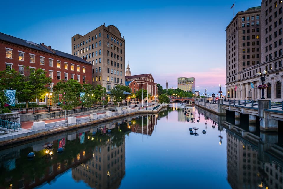 A calm river reflecting buildings, a bridge, and a clock tower at dusk.
