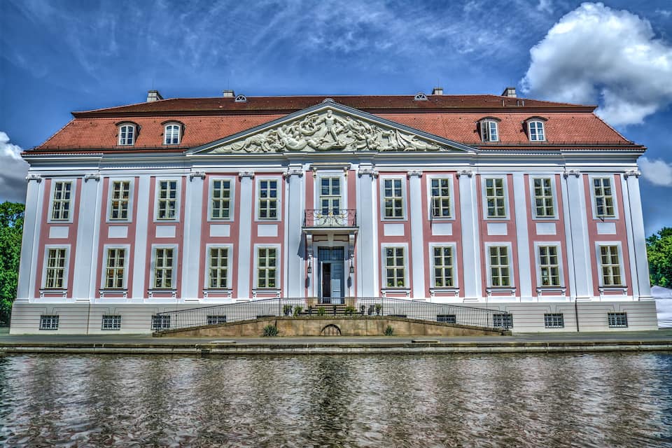 A large, historic building with a red roof and white and pink facade, situated by a calm body of water.
