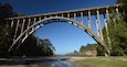 A large arched bridge over a river, surrounded by dense trees and a clear blue sky.