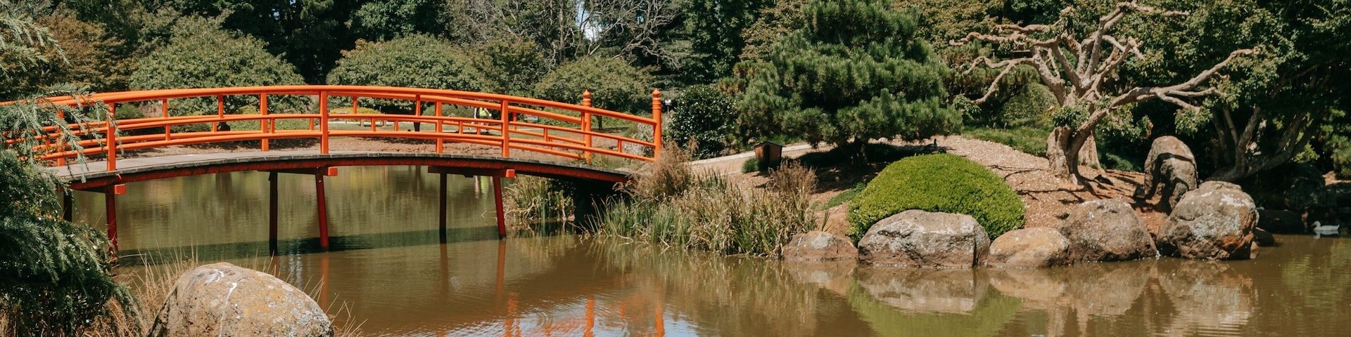 Uma ponte vermelha sobre um lago calmo, rodeada por árvores e pedras.