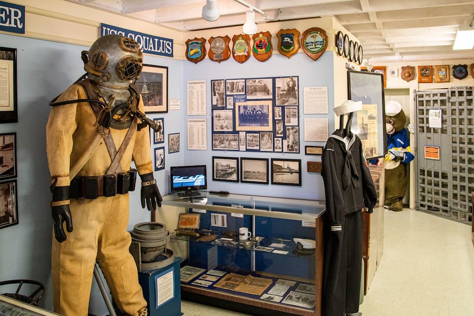 A museum exhibit with a life-sized submarine diver mannequin, historical photographs, and various medals and awards on display.