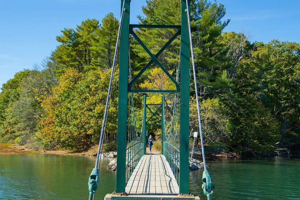 Un puente colgante de madera sobre un río tranquilo, rodeado de árboles con follaje otoñal.