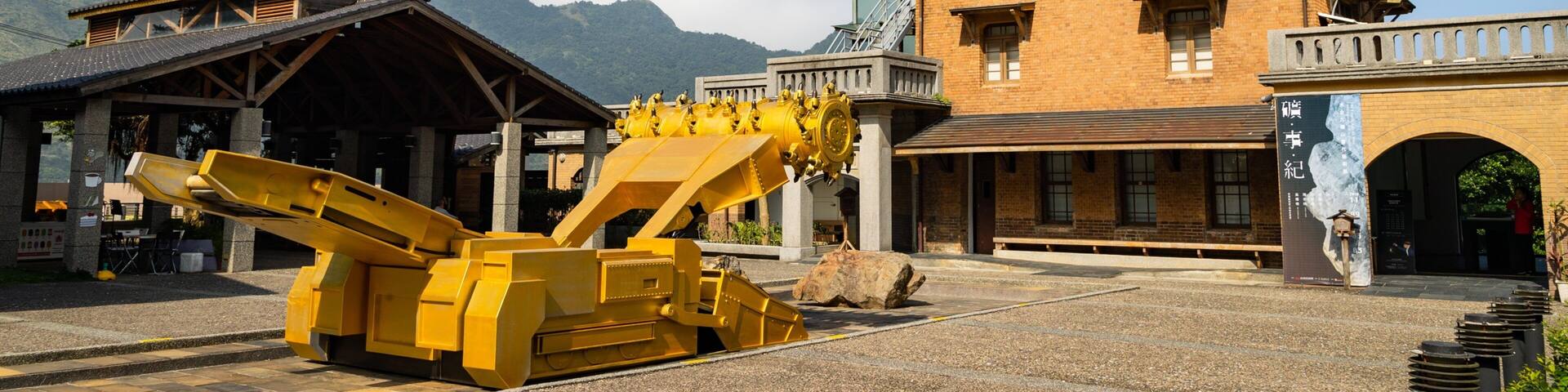 Una máquina minera amarilla grande en un patio con una montaña al fondo y un edificio con una torre del reloj.