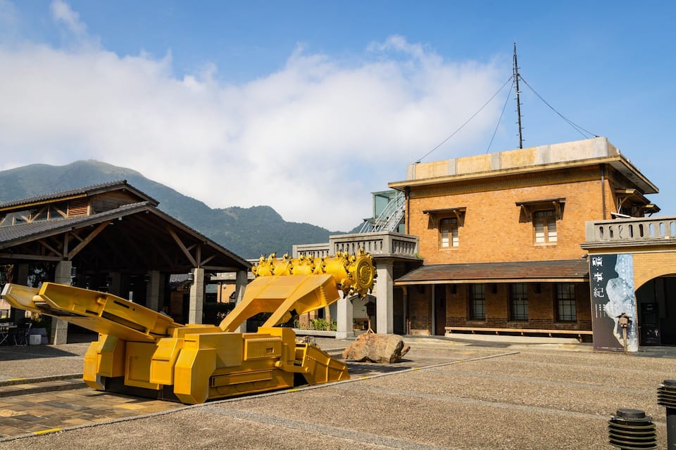 Una máquina minera amarilla grande en un patio con una montaña al fondo y un edificio con una torre del reloj.