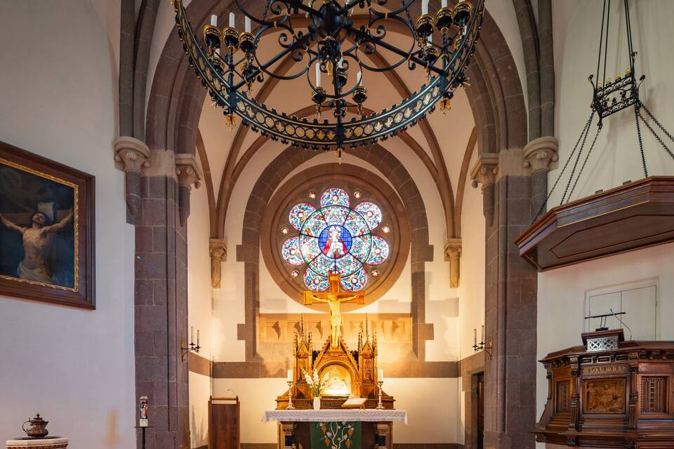 Interior de una iglesia con un amplio vitral, una cruz y un púlpito de madera.