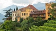 A historic building with a turret and a balcony, surrounded by lush greenery and mountains in the background.