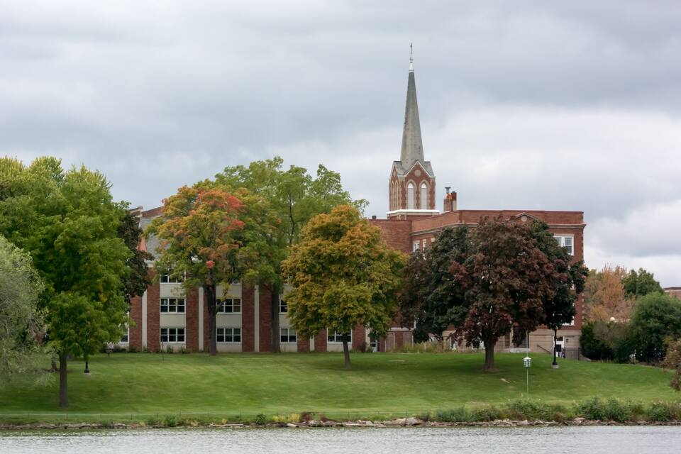 A church with a tall steeple, surrounded by trees and a body of water.