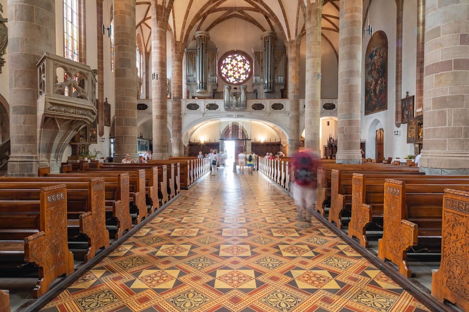 A church interior with ornate wooden pews, a patterned floor, and a large stained glass window.