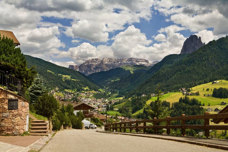 A mountain village with a stone building, wooden fence, and a road leading to a valley with houses and green hills.