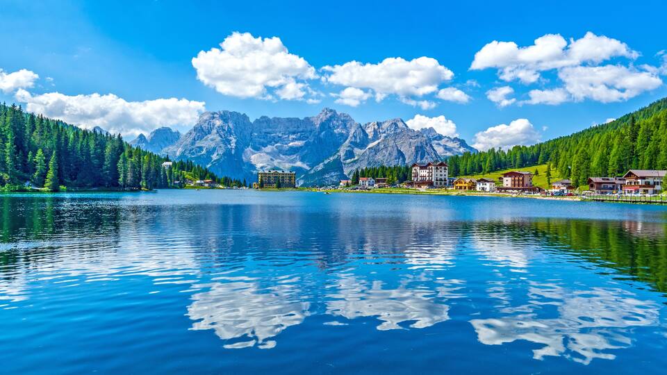 A calm lake with reflections, surrounded by mountains and a small town with buildings.