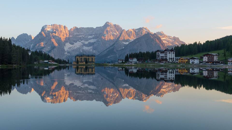 A mountain lake with reflections of buildings and trees, surrounded by forested hills and a prominent mountain range.