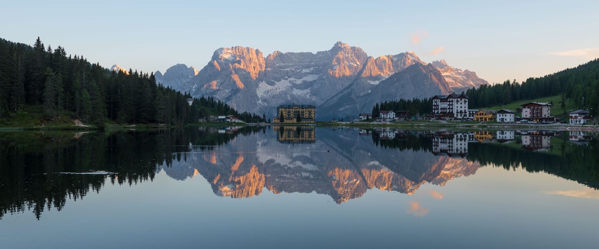 A mountain lake with reflections of buildings and trees, surrounded by forested hills and a prominent mountain range.