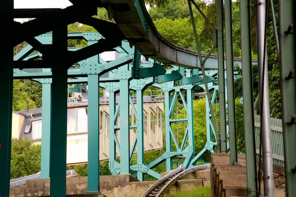 Un puente de metal verde con una vía curva, rodeado de vegetación y edificios residenciales.