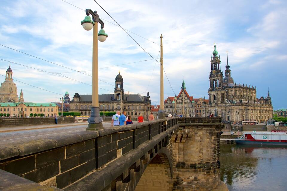 A historic bridge with stone railings, a river, and a large ornate church with multiple spires in the background.