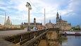Een historische brug met stenen hekwerk, een rivier en een grote, versierde kerk met meerdere torens op de achtergrond.
