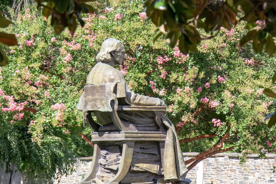 A statue of a seated man with a book, surrounded by flowering bushes and trees.