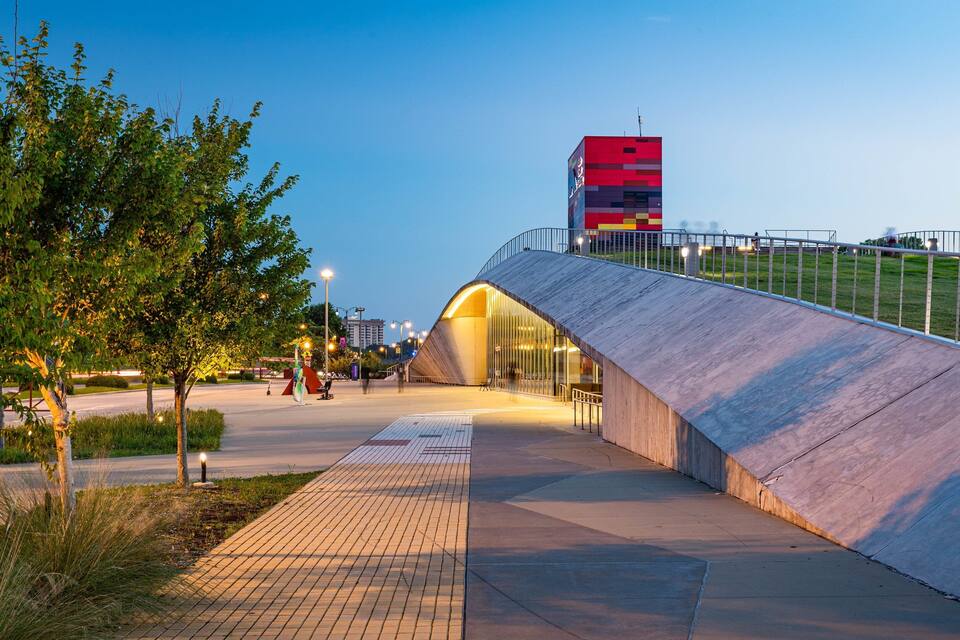 Un puente moderno de diseño curvo, barandilla metálica y un edificio con fachada roja y negra al fondo.