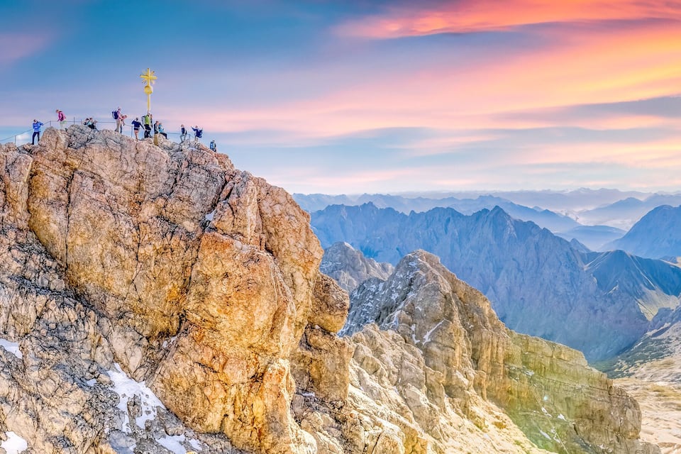 A rocky mountain peak with a golden cross at the summit, surrounded by rugged terrain and distant mountain ranges.